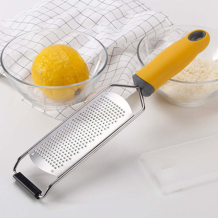 Stainless steel cheese grater with yellow handle, surrounded by sliced lemon and grated cheese in bowls, on a kitchen counter.