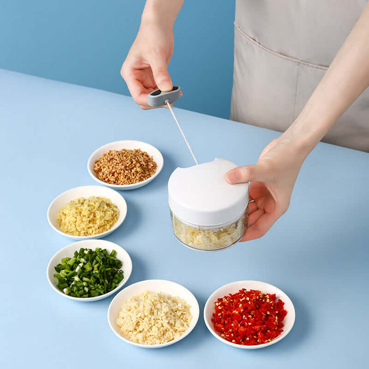 Manual garlic crusher in use with various chopped ingredients in bowls on a blue background.