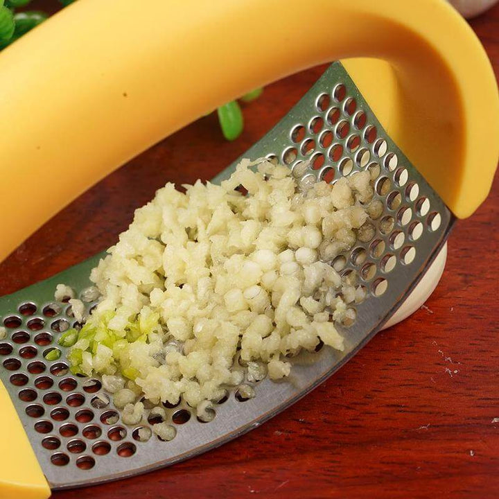 Close-up of a stainless steel garlic masher crushing fresh garlic on a wooden surface, highlighting its curved ergonomic design.