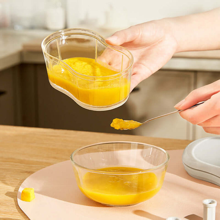 Person holding and spooning orange liquid from a curved glass container into a bowl on a kitchen counter.