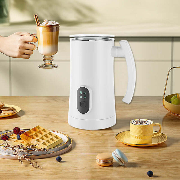 Automatic stainless steel electric milk frother on a kitchen countertop with waffles and coffee.