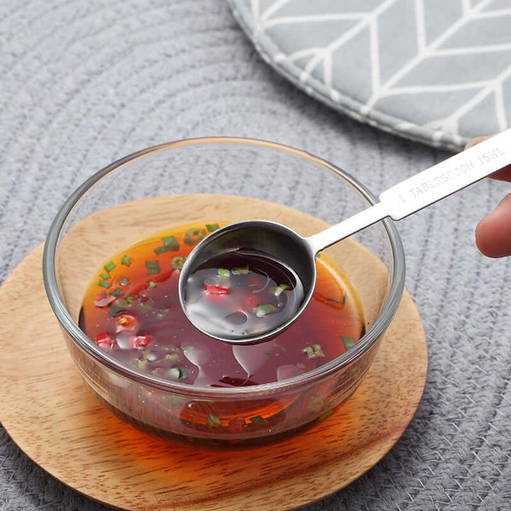 Stainless steel measuring spoon with liquid seasoning in glass bowl on wooden coaster.