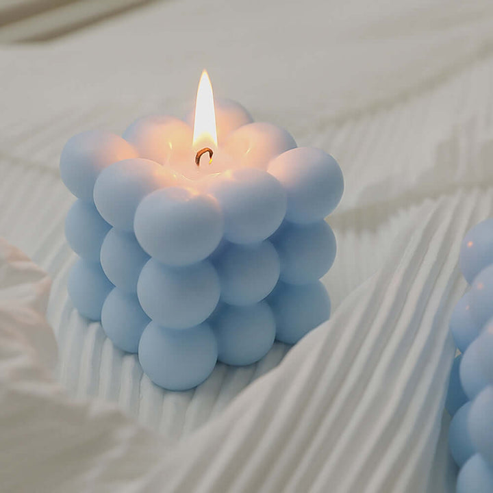 Teal geometric Rubik's Cube candle, lit, on textured white fabric for Japandi style decor.