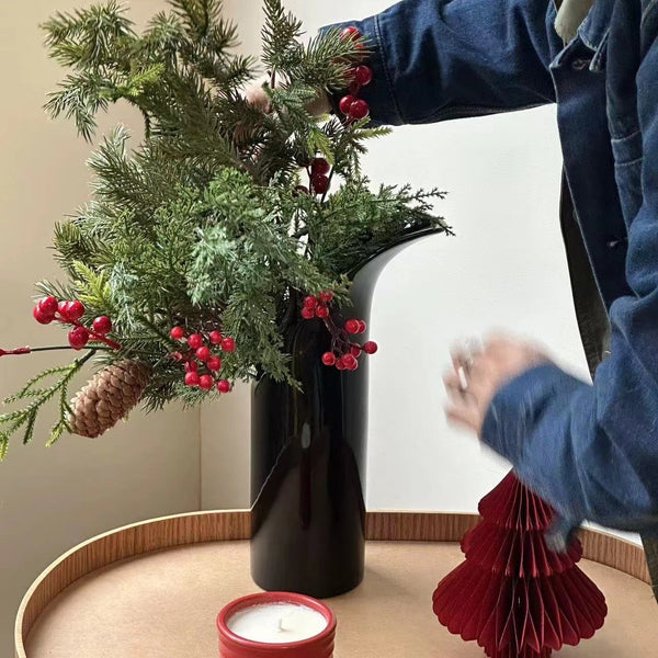 Person arranging festive greenery in a black vase on a table with candle.