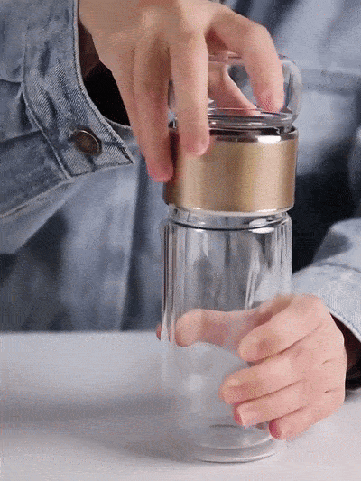 Hands demonstrating a glass tea infuser bottle with built-in stainless-steel filter and gold lid, ideal for home or travel use.
